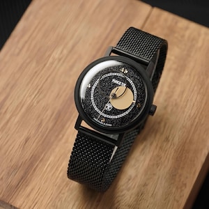May include: A black wristwatch with a textured mesh strap and a round case. The dial features a black speckled background, a gold circular accent, white hour markers, and the text "PAKETA" and "СДЕЛАНО В СССР" printed in white.