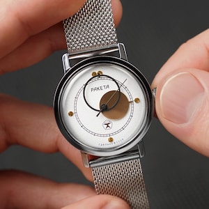 May include: A silver wristwatch with a mesh band. The watch face is white with a black ring, a gold-colored circle, and the word "RAKETA" in black. The watch is held by two hands.