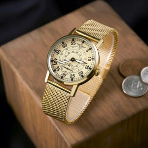 May include: A gold-toned wristwatch with a mesh band rests on a wooden block. The watch face is cream-colored with black numerals and hands. The watch has a small seconds dial and a logo at the top. The band is approximately 0.75 inches wide.