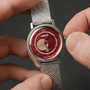 May include: A silver-tone wristwatch with a textured red dial, featuring a circular off-centre sub-dial, thin hands, and a mesh metal strap. The watch face displays the text PAKETA and СДЕЛАНО В СССР in white lettering with a small star emblem.