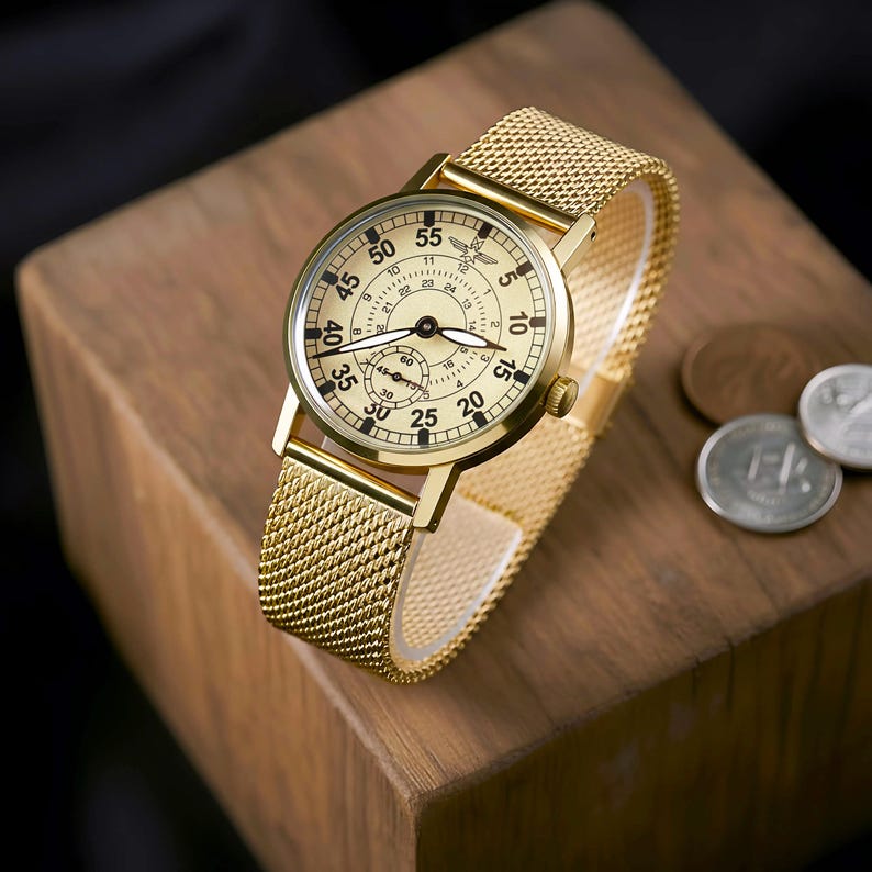 K&ouml;nnte beinhalten: Eine goldfarbene Armbanduhr mit einem Mesh-Armband liegt auf einem Holzblock. Das Zifferblatt der Uhr ist cremefarben mit schwarzen Ziffern und Zeigern. Die Uhr hat ein kleines Hilfszifferblatt und ein goldfarbenes Geh&auml;use. Der Durchmesser der Uhr betr&auml;gt ca. 3,8 cm.