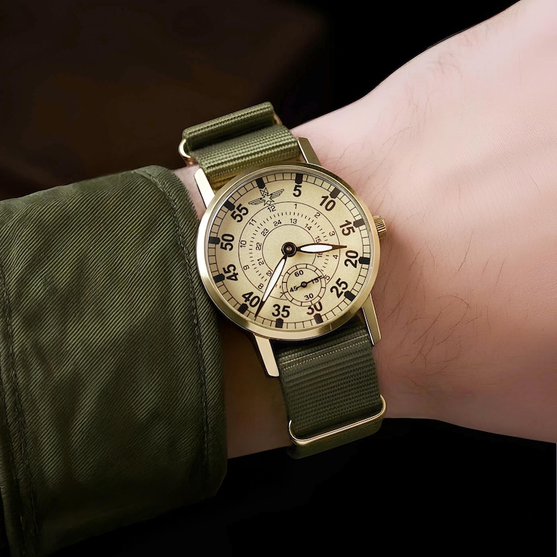 May include: A gold-toned wristwatch with a cream-colored face and a green nylon strap. The watch face features black numerals and a small subdial. The watch is secured on a wrist, showcasing its classic design and military-inspired aesthetic.
