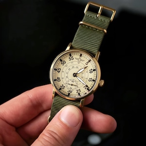 May include: A vintage-style wristwatch with a cream-colored face and gold-tone case. The watch features black numerals, a small seconds dial, and a green nylon strap with a gold buckle. The watch is held by a hand.