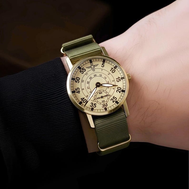 May include: A gold-toned wristwatch with a cream-colored face and black numerals. The watch has a green nylon strap and a small seconds dial. The watch face displays the numbers 5, 10, 15, 20, 25, 30, 35, 40, 45, 50, and 55.