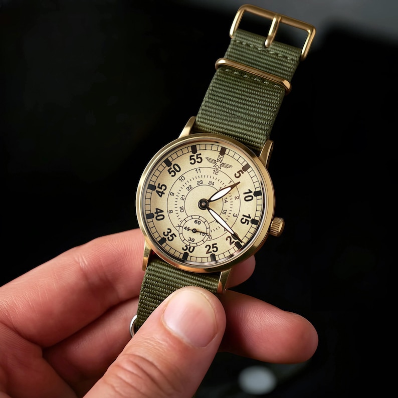 May include: A vintage-style wristwatch with a cream-colored face and black numerals. The watch has a gold-tone case and a green nylon strap. The watch face includes a small seconds dial and a 24-hour clock. The watch is held by a hand.