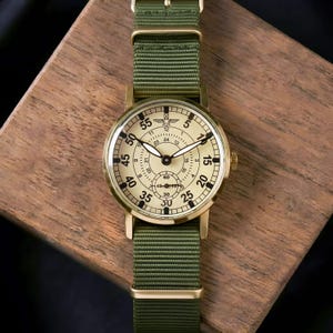 May include: A vintage-style wristwatch with a gold-colored case and a cream-colored face. The watch features black numerals and hands, with a smaller dial at the bottom. It has a green nylon strap and is set on a wooden surface.