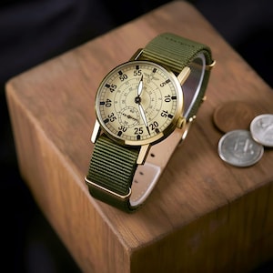 May include: A gold-toned wristwatch with a cream-colored face and black numerals, featuring a green nylon band. The watch has a subdial and is set on a wooden block. The watch face displays the numbers 5, 10, 15, 20, 25, 30, 35, 40, 45, 50, and 55.