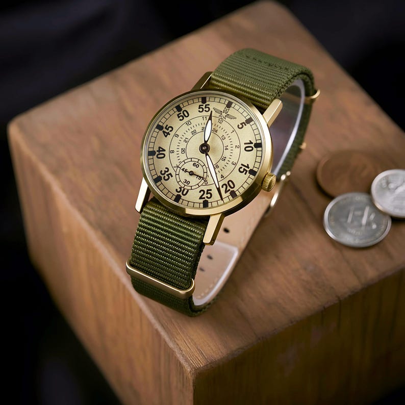 May include: A gold-toned wristwatch with a cream-colored face and black numerals, featuring a green nylon band. The watch has a subdial and is set on a wooden block. The watch face displays the numbers 5, 10, 15, 20, 25, 30, 35, 40, 45, 50, and 55.