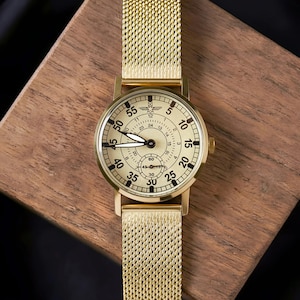 May include: A gold-toned wristwatch with a mesh band, set against a wooden surface. The watch face is cream-colored with black numerals and hands. The watch has a small seconds dial and a crown on the side.