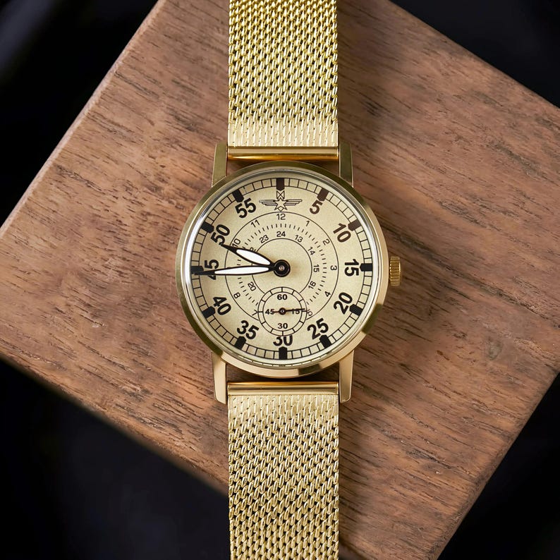 May include: A gold-toned wristwatch with a mesh band, set against a wooden surface. The watch face is cream-colored with black numerals and hands. The watch has a small seconds dial and a crown on the side.