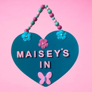 May include: Teal heart-shaped sign with the name "Maisey's In" written in pink letters. The sign is decorated with pink and blue flowers and a butterfly.
