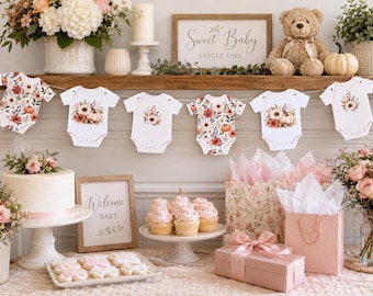 May include: A baby shower display featuring a banner of six white baby bodysuits with floral and pumpkin prints, a cake on a stand, cupcakes, gift bags, a pink gift box, and framed signs reading Sweet Baby Little One and Welcome Baby.