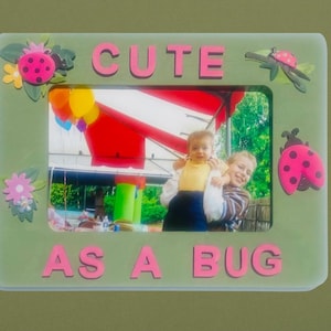 Puede incluir: Un marco de fotos verde con un borde rosa y decoraciones de mariquitas. El marco tiene las palabras "Cute as a Bug" escritas en letras rosas. El marco contiene una foto de dos niños, uno sosteniendo al otro.