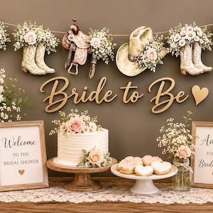 May include: A bridal shower display featuring a Bride to Be sign, a white cake on a wooden stand, heart-shaped cookies, two framed signs, and floral arrangements. A banner above shows cowboy boots, a saddle, a hat, and a cactus with flowers.