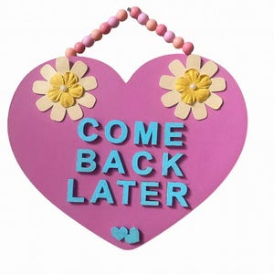 May include: Pink heart-shaped wooden sign with a beaded hanger. The sign says "COME BACK LATER" in blue letters. The sign is decorated with two yellow flowers with white petals.