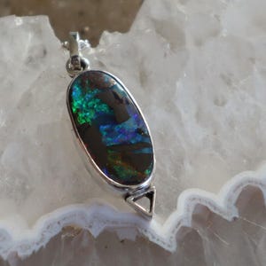 May include: A silver pendant with an oval-shaped black opal. The opal has iridescent green and blue flashes. The pendant has a silver bezel and a triangular bail. The opal is approximately 1 inch long.