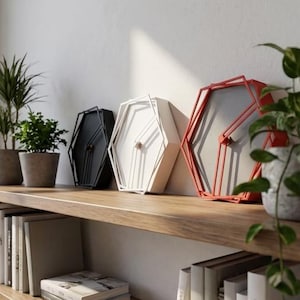 May include: Three hexagonal wall clocks in black, white, and red. Each clock features a geometric wire frame design with a central dial and hands. The clocks are arranged in a row, showcasing their distinct colors and angular, open-frame construction.