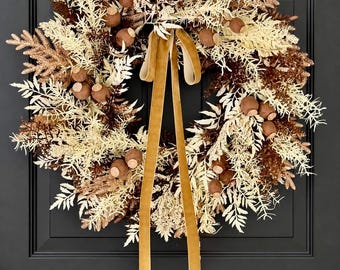 May include: A circular wreath featuring a mix of cream-colored and dark brown foliage, accented with small, round, brown seed pods. A long, smooth, golden-brown velvet ribbon hangs from the top center, extending downward in two long tails.