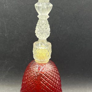 May include: A red glass bell-shaped bottle with a clear glass stopper. The bottle has a diamond-shaped pattern and the stopper has a multi-tiered design.