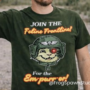 Può includere: T-shirt verde scuro con la scritta "JOIN THE Feline Frontline!" e "For the Em-purr-or!" raffigurante un gatto con un elmetto militare. La persona nell'immagine indossa un berretto mimetico e occhiali da sole.