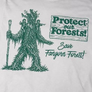 Op de afbeelding: Wit t-shirt met een groene afbeelding van een boomachtig figuur met een staf. De tekst "Protect our Forests! Save Fangorn Forest!" staat op het t-shirt. Het t-shirt wordt getoond op een mosachtig oppervlak.