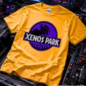 May include: A mustard yellow short-sleeved t-shirt featuring a circular purple graphic with a silhouette of a creature and the text XENOS PARK in white letters. The shirt is displayed against a dark, detailed cockpit control panel background.