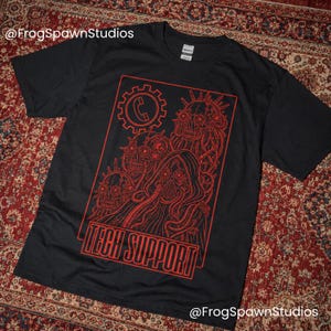 Op de afbeelding: Een zwart t-shirt met korte mouwen en een rode grafische print van drie mechanische schedels en een figuur met een capuchon en kabels. Boven de figuren staat een telefoonicoon in een tandwiel. De tekst TECH SUPPORT staat onderaan in het rood.