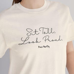 Può includere: Una maglietta bianca con la scritta "Sit Tall, Look Proud. Panic Quietly" stampata sul davanti.