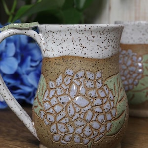 May include: Handmade ceramic mug with a white speckled rim and handle. The mug features a brown band with a blue and green floral design. The handle has a green accent. A second mug is visible in the background.