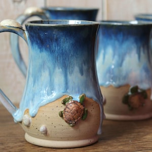 May include: Ceramic mugs with a blue and white glaze, resembling ocean waves. Each mug features a sculpted sea turtle and seashells. The mugs have a light blue handle and a beige base, creating a coastal theme.