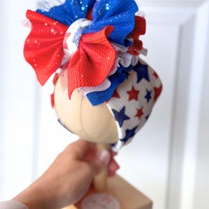 May include: A patriotic red, white, and blue baby headband with a large bow. The bow features layers of red and blue fabric with glitter accents and a white star-patterned band. The headband is displayed on a wooden stand.