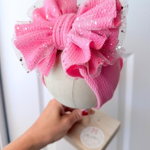 May include: A pink baby headband with a large bow. The bow is made of textured fabric and has a layer of sparkly, sheer material on top. The headband is displayed on a mannequin head.