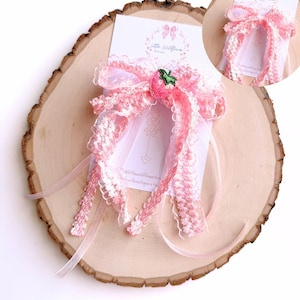 May include: A pink and white hair bow with a strawberry accent, displayed on a wooden slice. The bow features lace trim and sheer ribbons. A small card with the text "Little Wildflower Boutique" is visible.