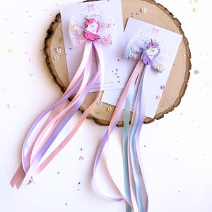 May include: Two unicorn hair bows with long, pastel-colored ribbons. Each bow features a unicorn head and is attached to a white card with the text "Little Wildflowers". The bows are displayed on a wooden slice, with scattered star-shaped confetti.