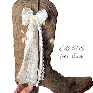 May include: A brown leather cowboy boot adorned with a white lace bow and cream-colored trim. The boot has decorative stitching and metal studs. Text on the right reads "Kids + Adult Lace Bows".
