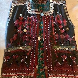 May include: A dark brown vest with intricate embroidery and beading. The vest has a high neckline and is adorned with silver buttons and white beads. The vest is made of a thick fabric and has a traditional design.
