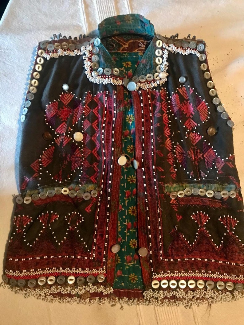 May include: A dark brown vest with intricate embroidery and beading. The vest has a high neckline and is adorned with silver buttons and white beads. The vest is made of a thick fabric and has a traditional design.
