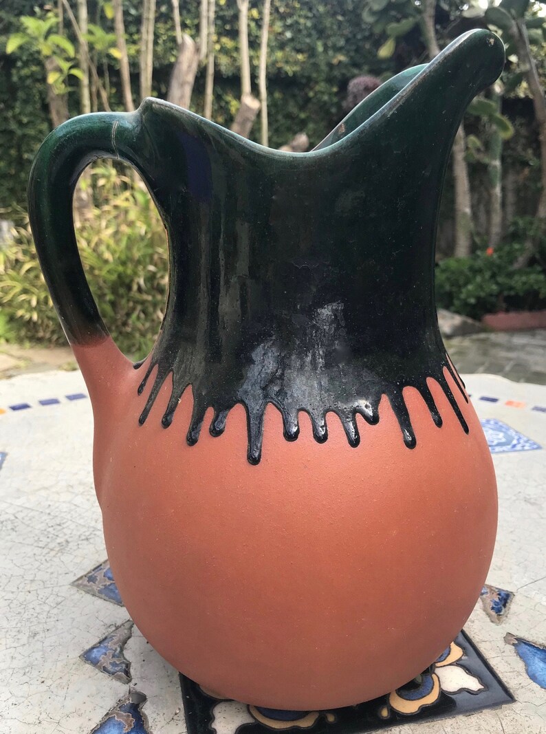 May include: A terracotta pitcher with a black and green glaze. The glaze has a drip effect around the top of the pitcher.