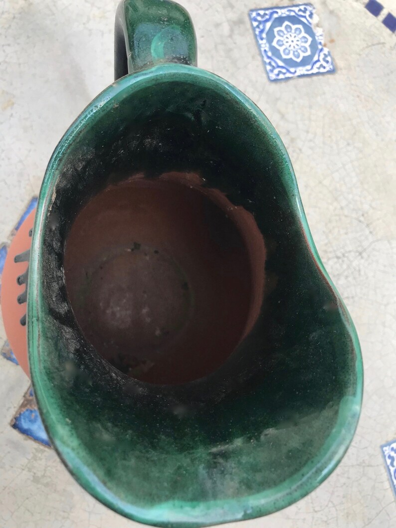 May include: A green glazed ceramic pitcher with a brown clay interior. The pitcher has a wide mouth and a handle.