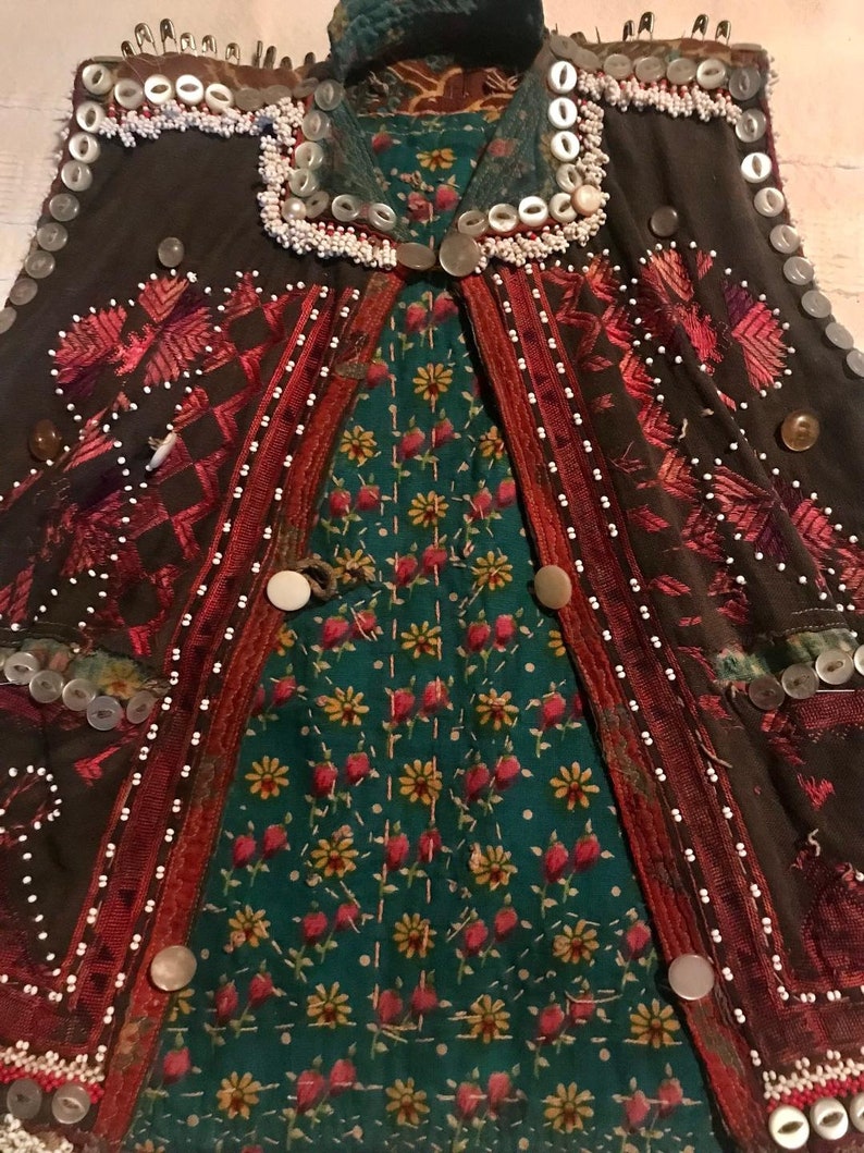 May include: A brown vest with intricate embroidery and beading. The vest is decorated with buttons, safety pins, and white beads. The vest has a green and red floral lining.