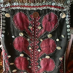 May include: A black vest with red embroidery and white beads. The vest is decorated with white shell buttons and a silver chain.