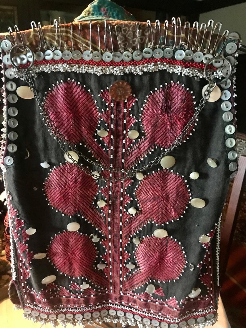 May include: A black vest with red embroidery and white beads. The vest is decorated with white shell buttons and a silver chain.