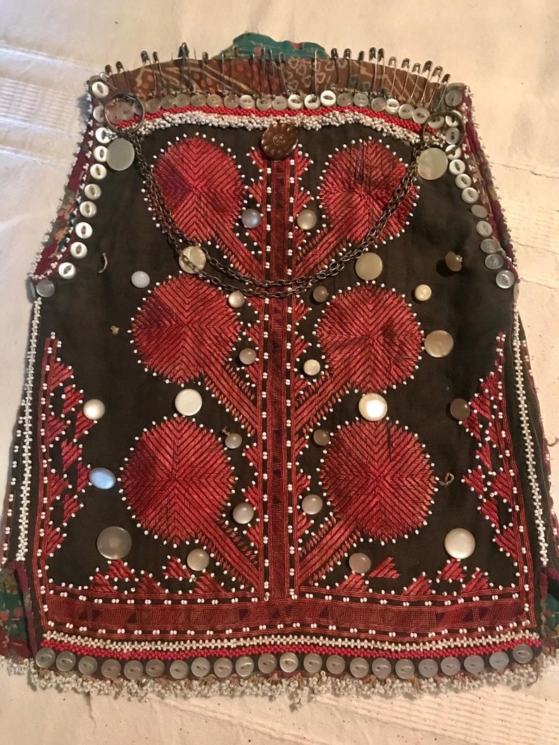 May include: A brown vest with red embroidery and white beads. The vest is decorated with silver buttons and a chain with silver charms.