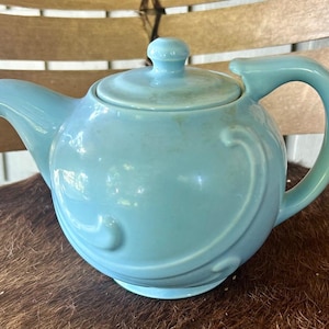 May include: A light blue ceramic teapot with a lid and handle. The teapot has a curved spout and a decorative swirl design on the body. The teapot is sitting on a brown surface.