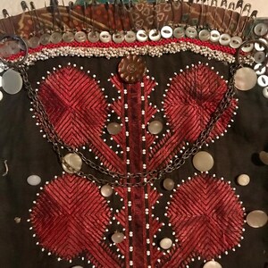 May include: A dark brown fabric garment with red embroidered designs and white beads. The garment is decorated with a chain and several buttons. The top of the garment is secured with safety pins.