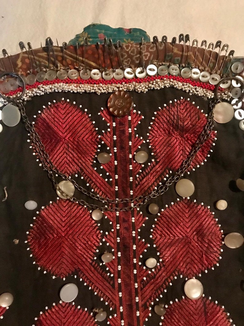 May include: A dark brown fabric garment with red embroidered designs and white beads. The garment is decorated with a chain and several buttons. The top of the garment is secured with safety pins.