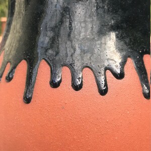 May include: A terracotta vase with a black glaze that drips down the sides. The glaze is thick and glossy, and the vase has a smooth, rounded shape.