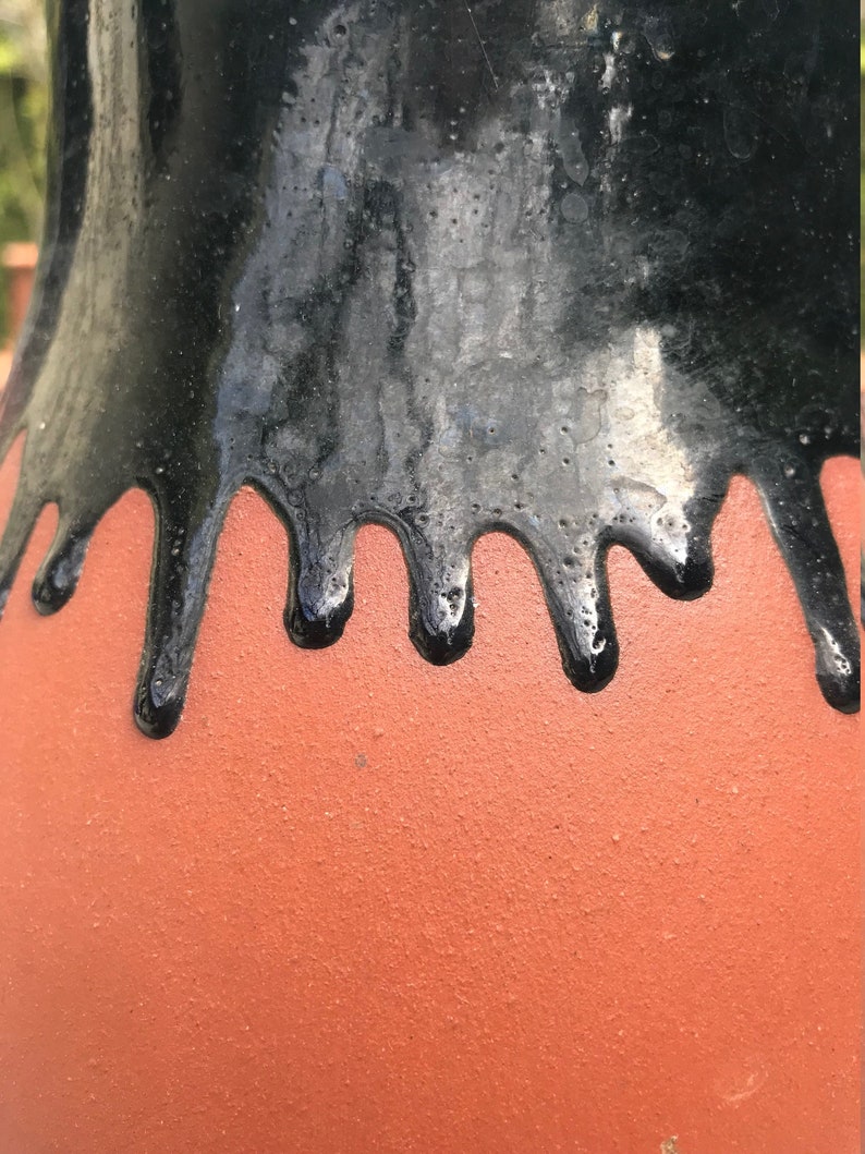 May include: A terracotta vase with a black glaze that drips down the sides. The glaze is thick and glossy, and the vase has a smooth, rounded shape.