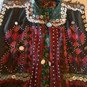 May include: A dark brown vest with intricate embroidery in shades of pink, red, and white. The vest is adorned with a variety of buttons, including white, gray, and brown. The vest has a collar and a front closure.