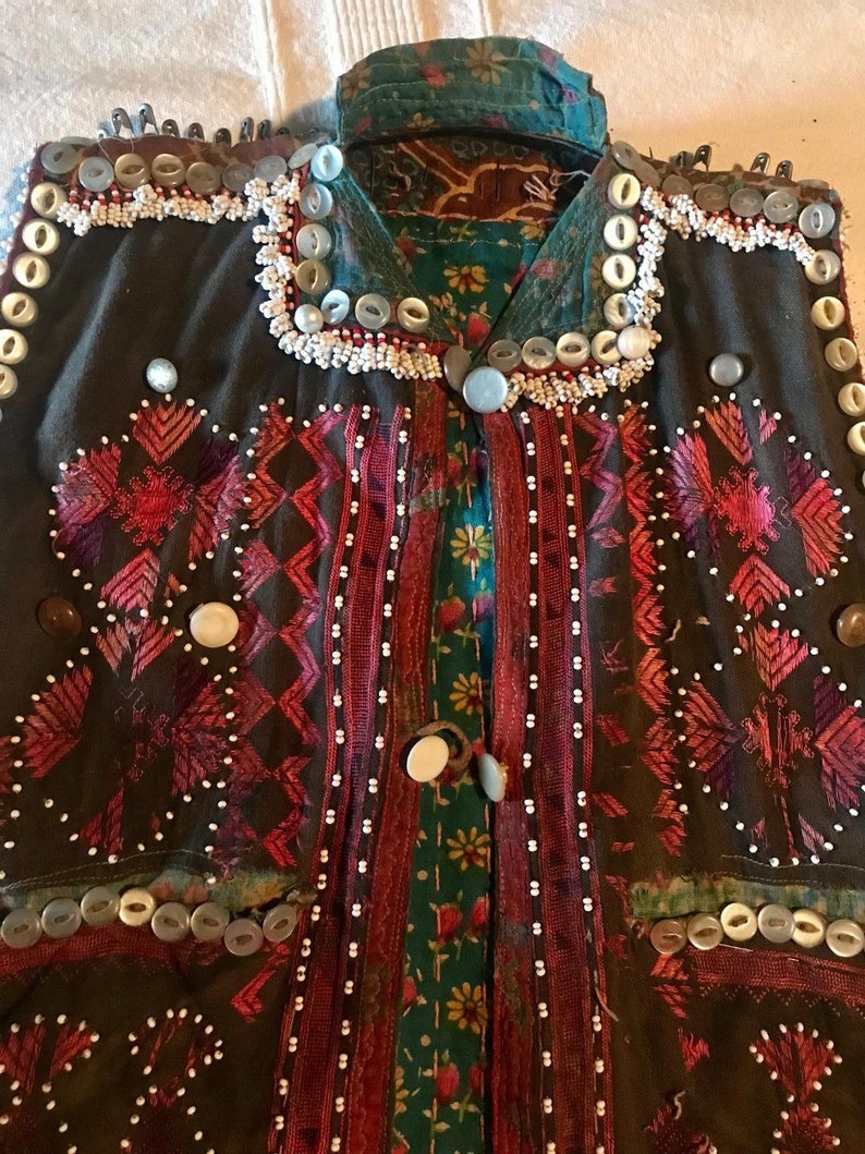May include: A dark brown vest with intricate embroidery in shades of pink, red, and white. The vest is adorned with a variety of buttons, including white, gray, and brown. The vest has a collar and a front closure.